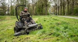 Grondverzorger bedient een zero-turn maaier op een sportterrein omgeven door bomen en gras.
