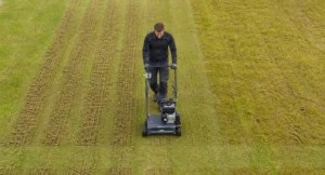Man verticuteert gazon met een verticuteerder, waarbij duidelijk zichtbaar is hoe het dode mos en strooisel wordt verwijderd.